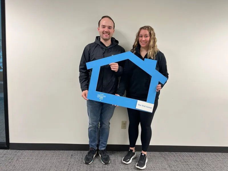 Two people holding a blue house-shaped cutout, standing in front of a white wall.