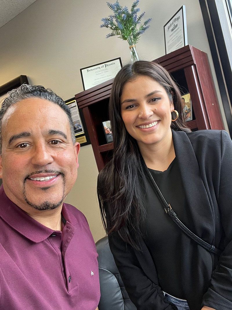 Two people smiling, indoors. Man in burgundy shirt, woman in black blazer. Wooden cabinet in background.