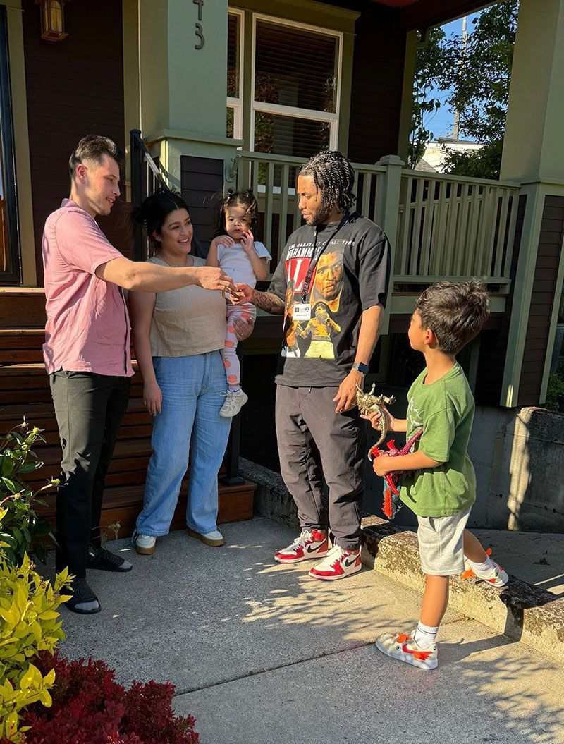 People exchanging keys in front of a house. Two adults and a child on the porch. A small child hands flowers.