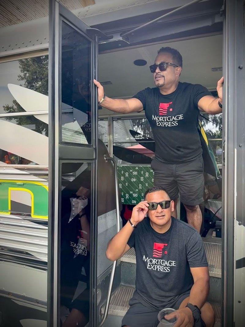 Two men wearing sunglasses and matching shirts stand in a doorway. One adjusts his glasses.