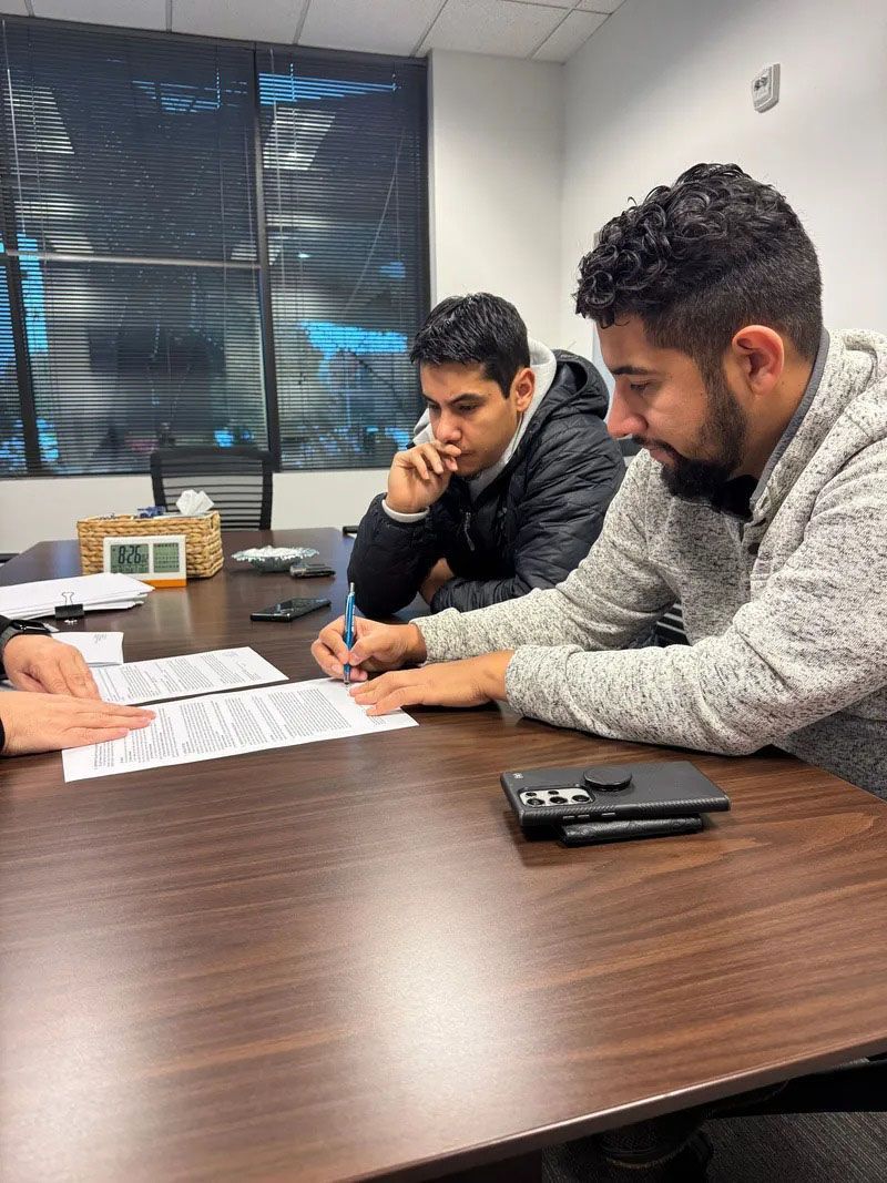 Two men reviewing documents at a table; one is writing. Interior office setting.