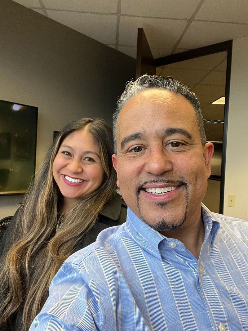 Two people smiling, indoors. Man in blue shirt takes selfie with woman with long hair.