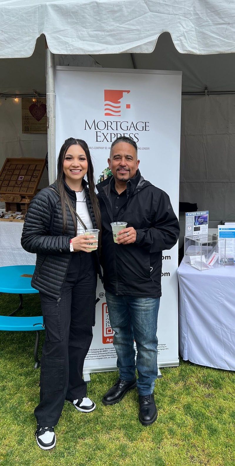 Two people stand near a banner for Mortgage Experts. Woman in black jacket, pants, and sneakers; man in black jacket and jeans. Both hold drinks.