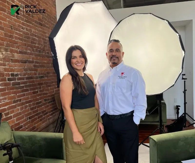 Woman and man pose for photo in a studio with softbox lights. Brick wall, green chairs. Man wears logo shirt.