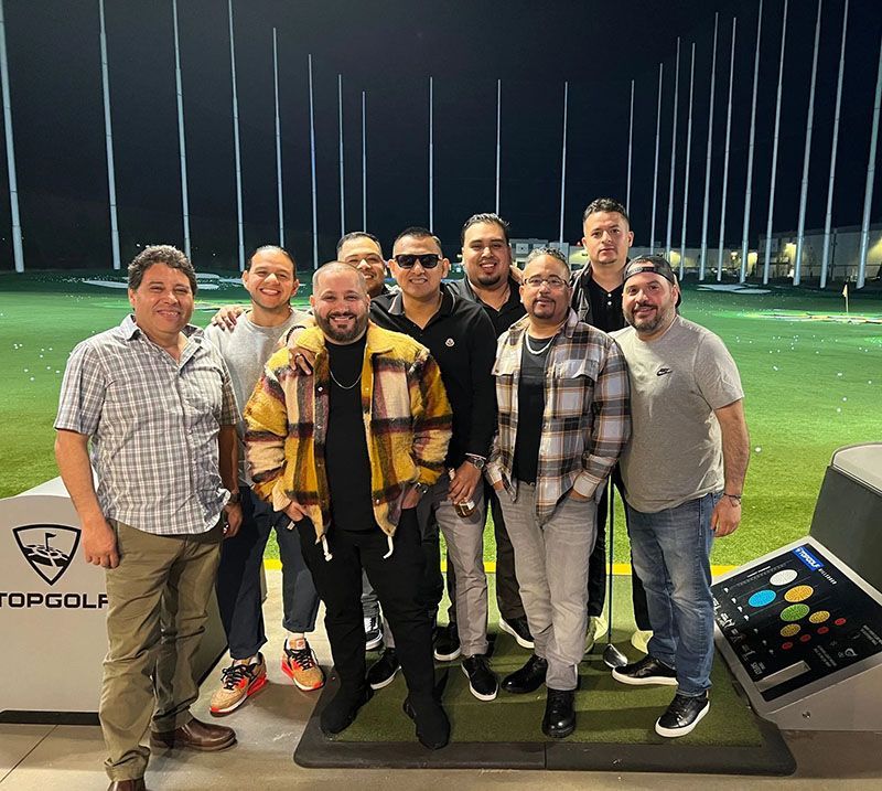 Group of men at Topgolf, posing near hitting stalls at night.