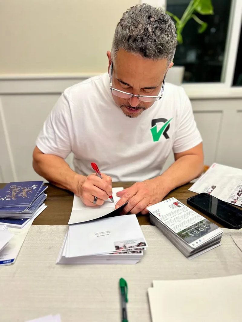 Man in white shirt with green logo, writing at a table with envelopes, papers, and pen.