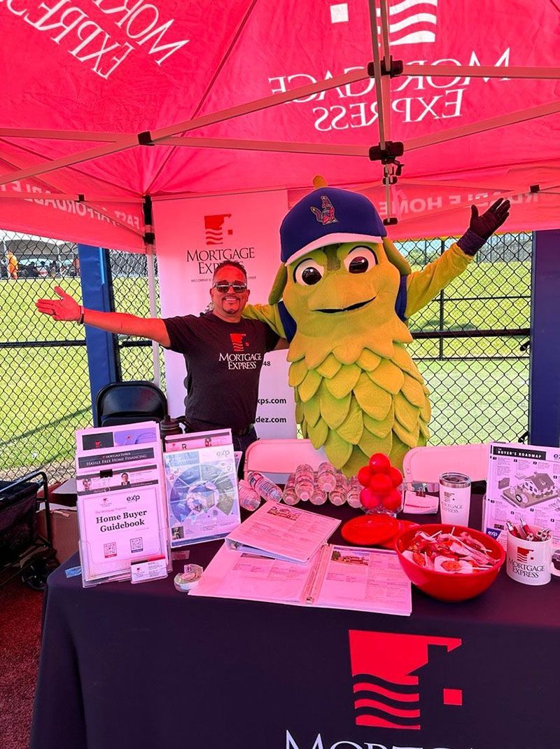 Man and mascot at a booth under a tent. They have arms open and smiling at the camera.