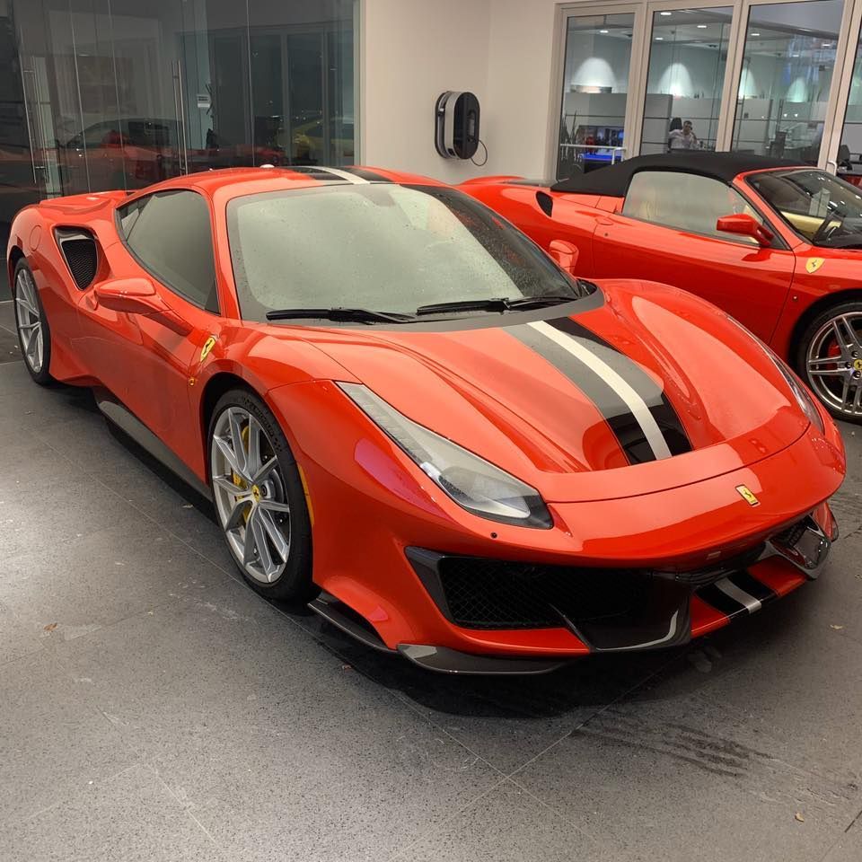 Red Ferrari sports car with black racing stripes in a showroom, another red car in the background.