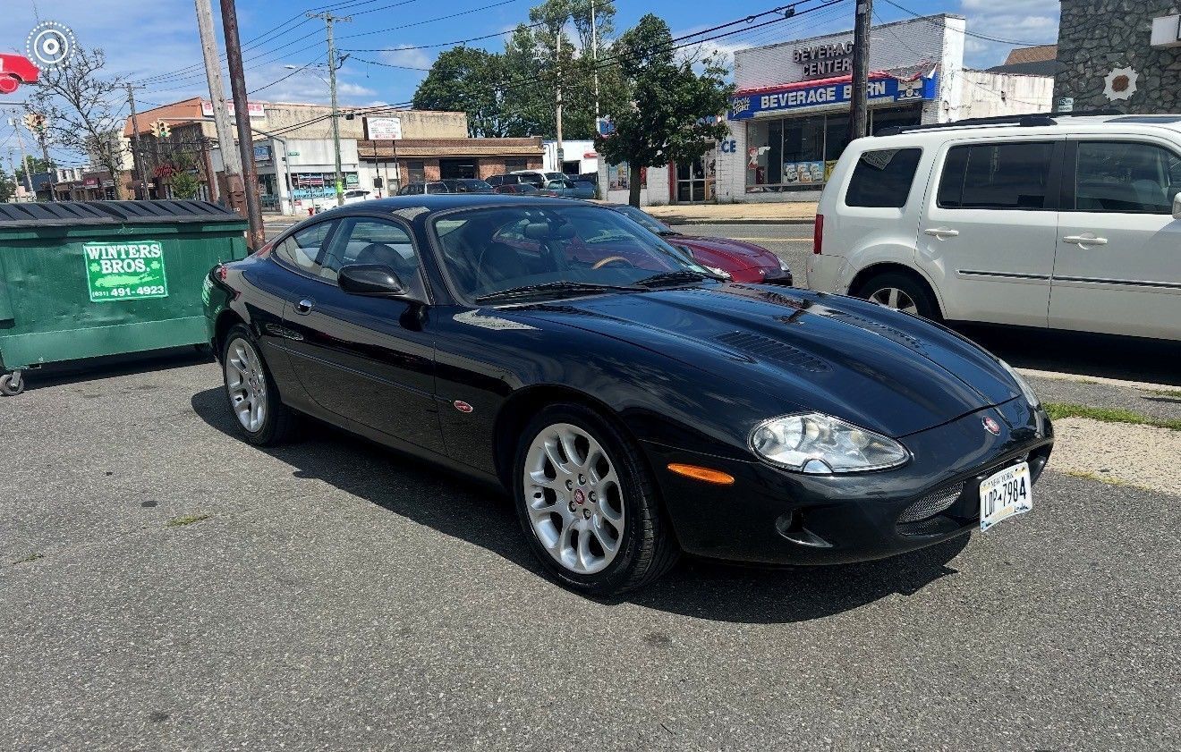 Black Jaguar coupe parked on asphalt, with a green dumpster and white SUV in the background.