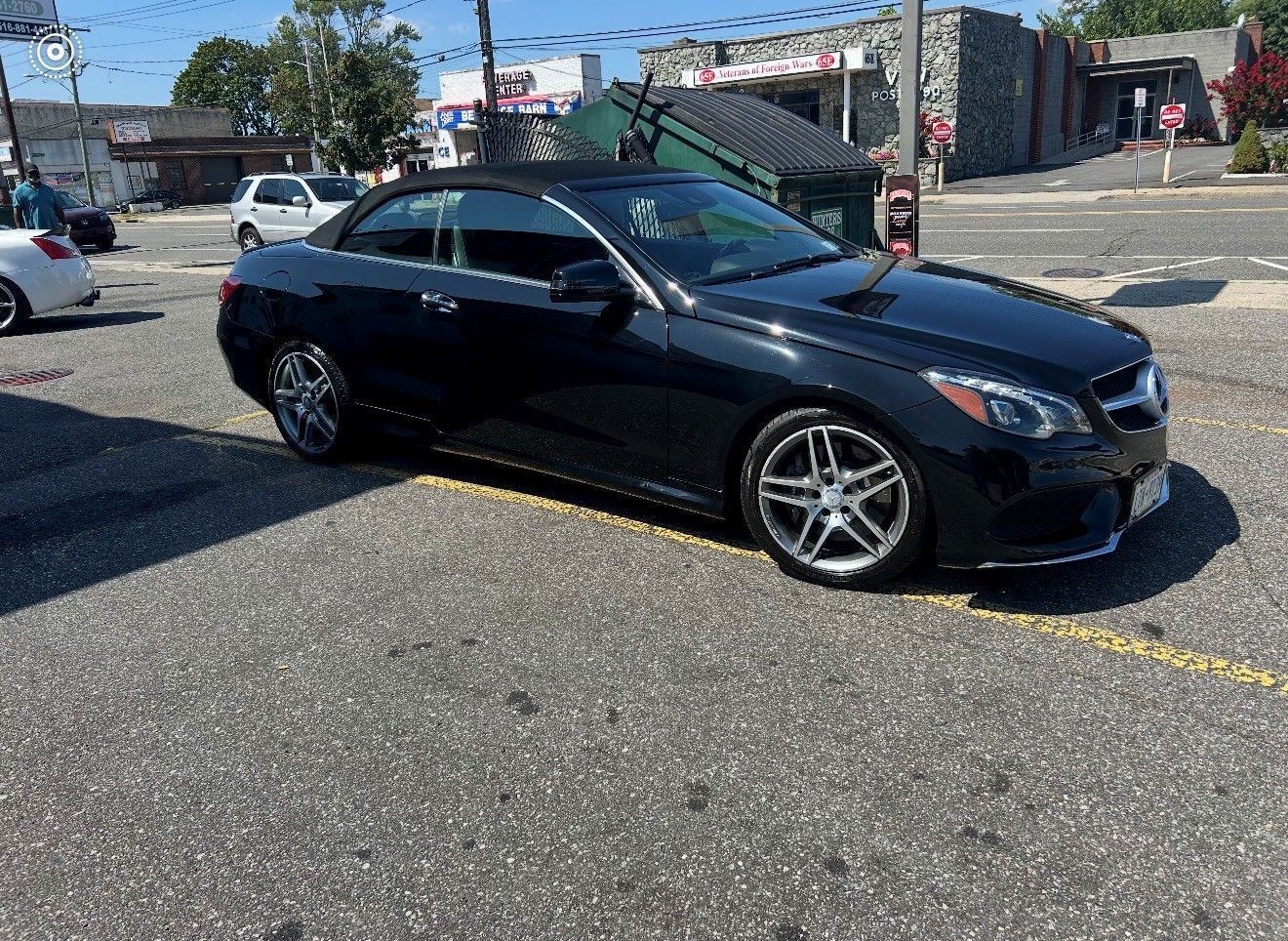 Black convertible car parked on asphalt road in front of storefronts on a sunny day.