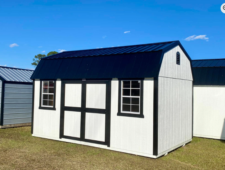 White and black barn-style shed with a black roof and two windows. Set on grass with a blue sky.