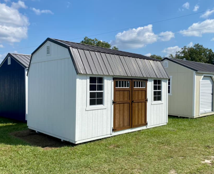 White barn-style shed with brown doors, metal roof, and two windows. Other sheds are in the background on grass.
