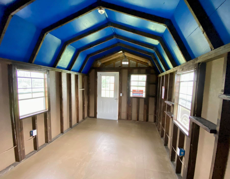 Interior of a shed with blue vaulted ceiling, windows, and a white door. Brown framing and tan flooring.