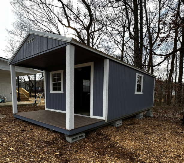Blue shed with porch, white trim, on concrete blocks in wooded area.
