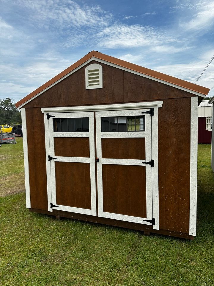 Brown shed with white trim, double doors, and a small vent under a tan roof, set on grass.