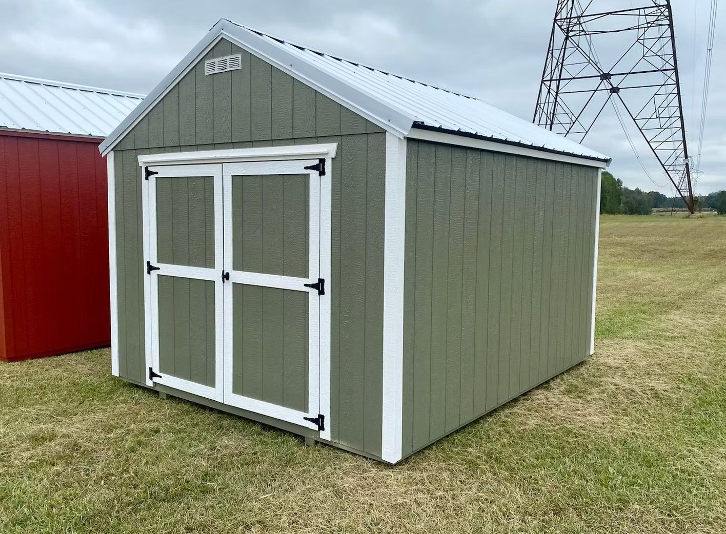 Green shed with white trim, double doors, and metal roof, sits in grassy field.