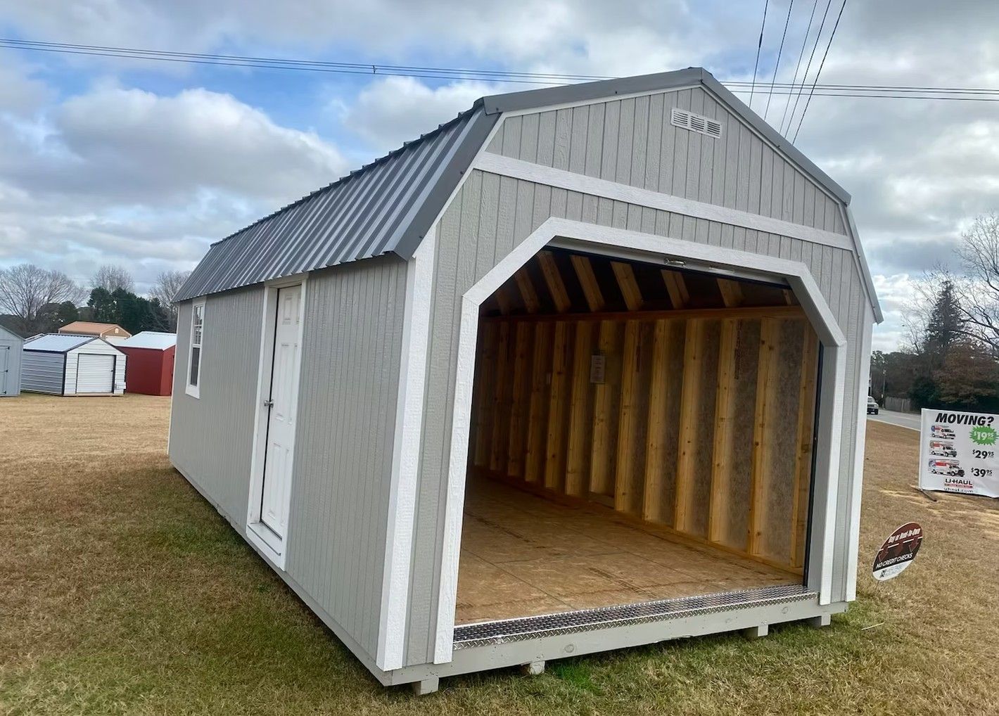 Gray barn-style shed with a large open doorway, white trim, and a metal roof on a grassy field.