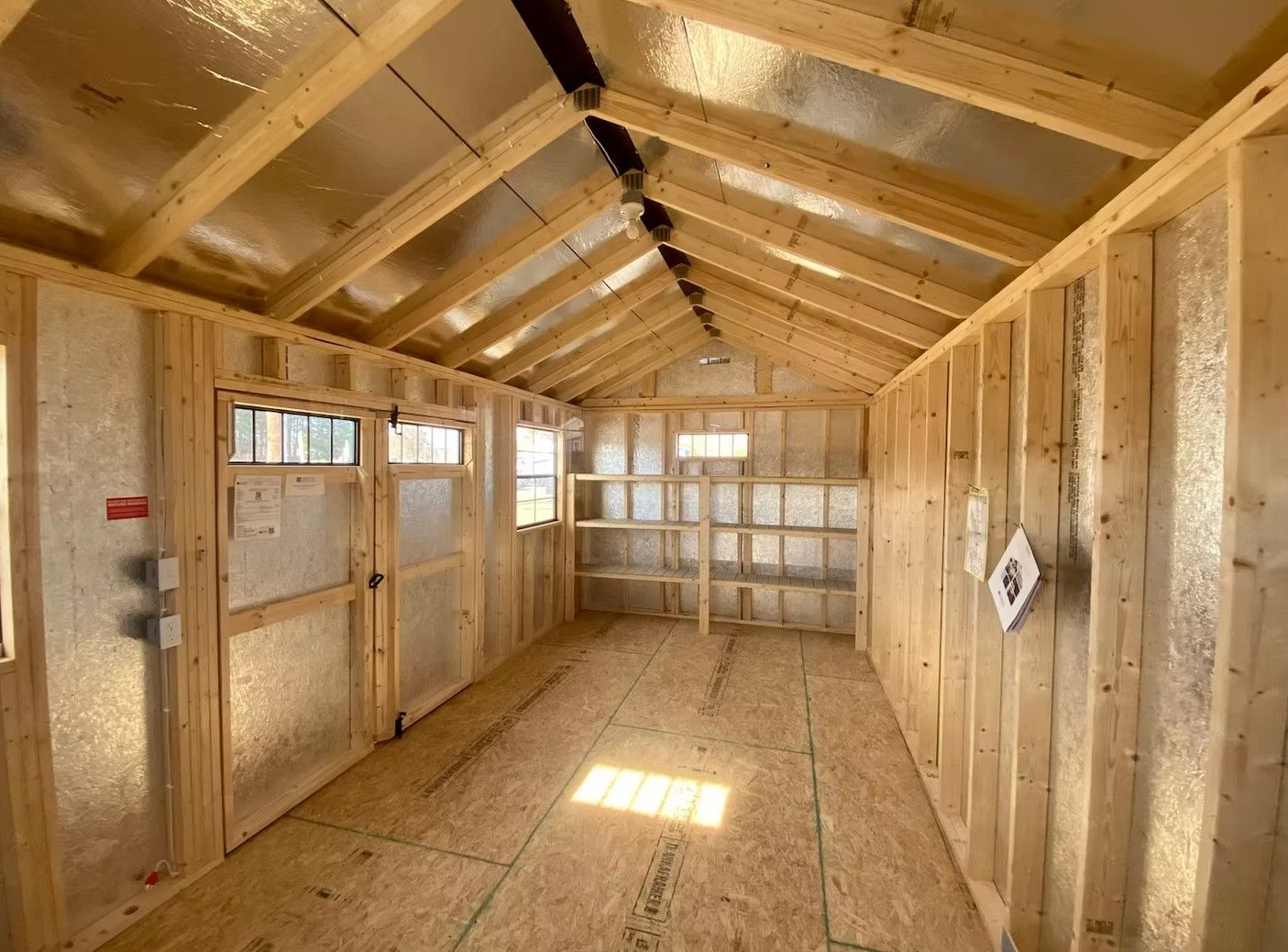 Interior of a wooden shed under construction, with open door, windows, and shelving.