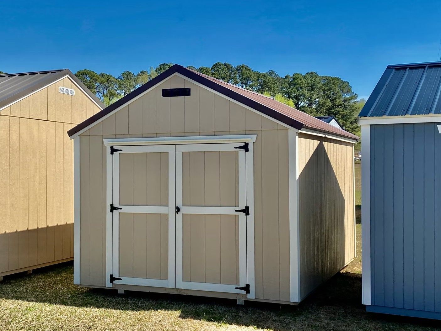 Tan shed with brown roof and white-trimmed double doors, outdoors on grass, with blue sky.