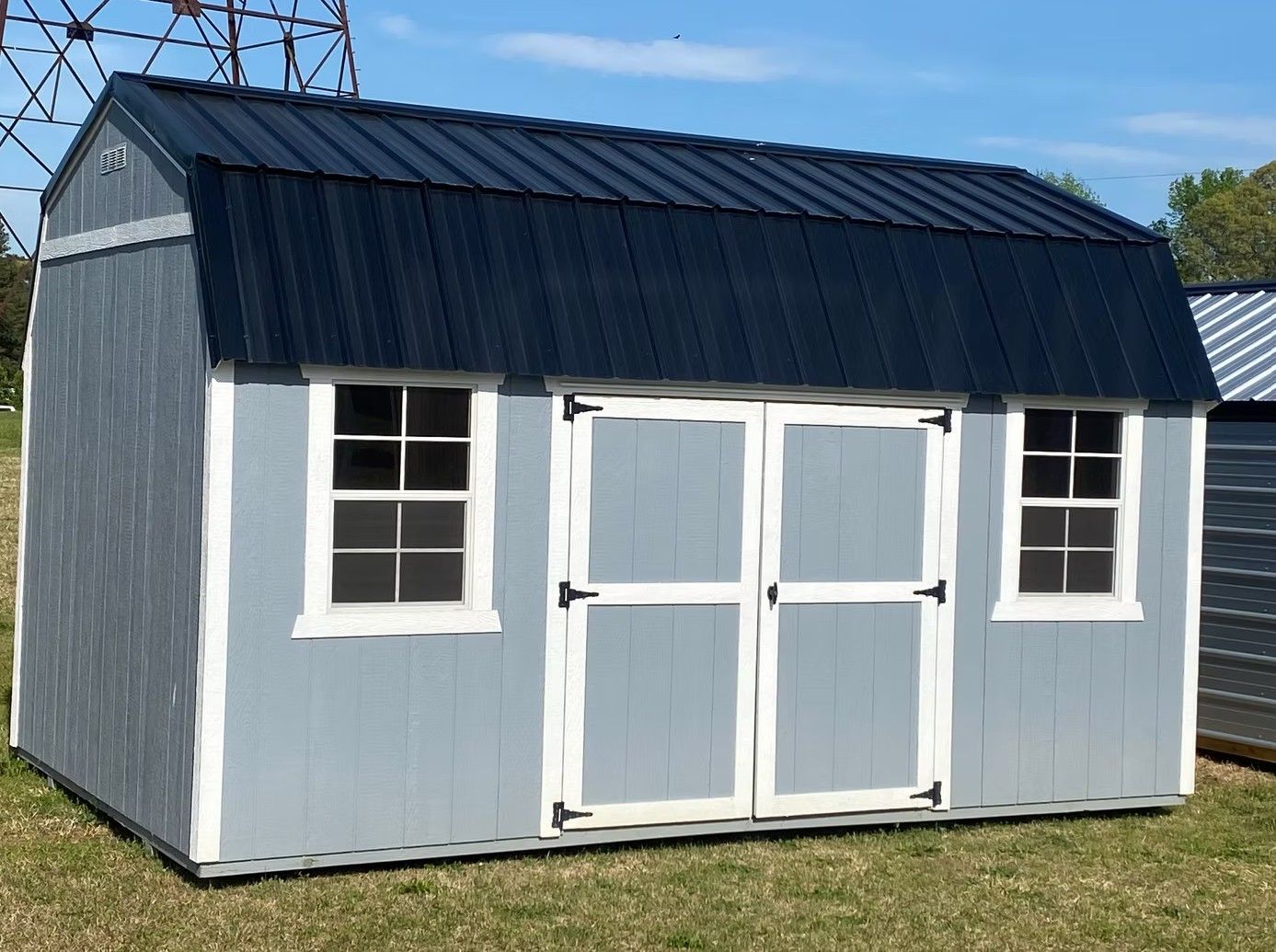 Light blue barn shed with white trim, black roof, two windows and double doors, on grass.
