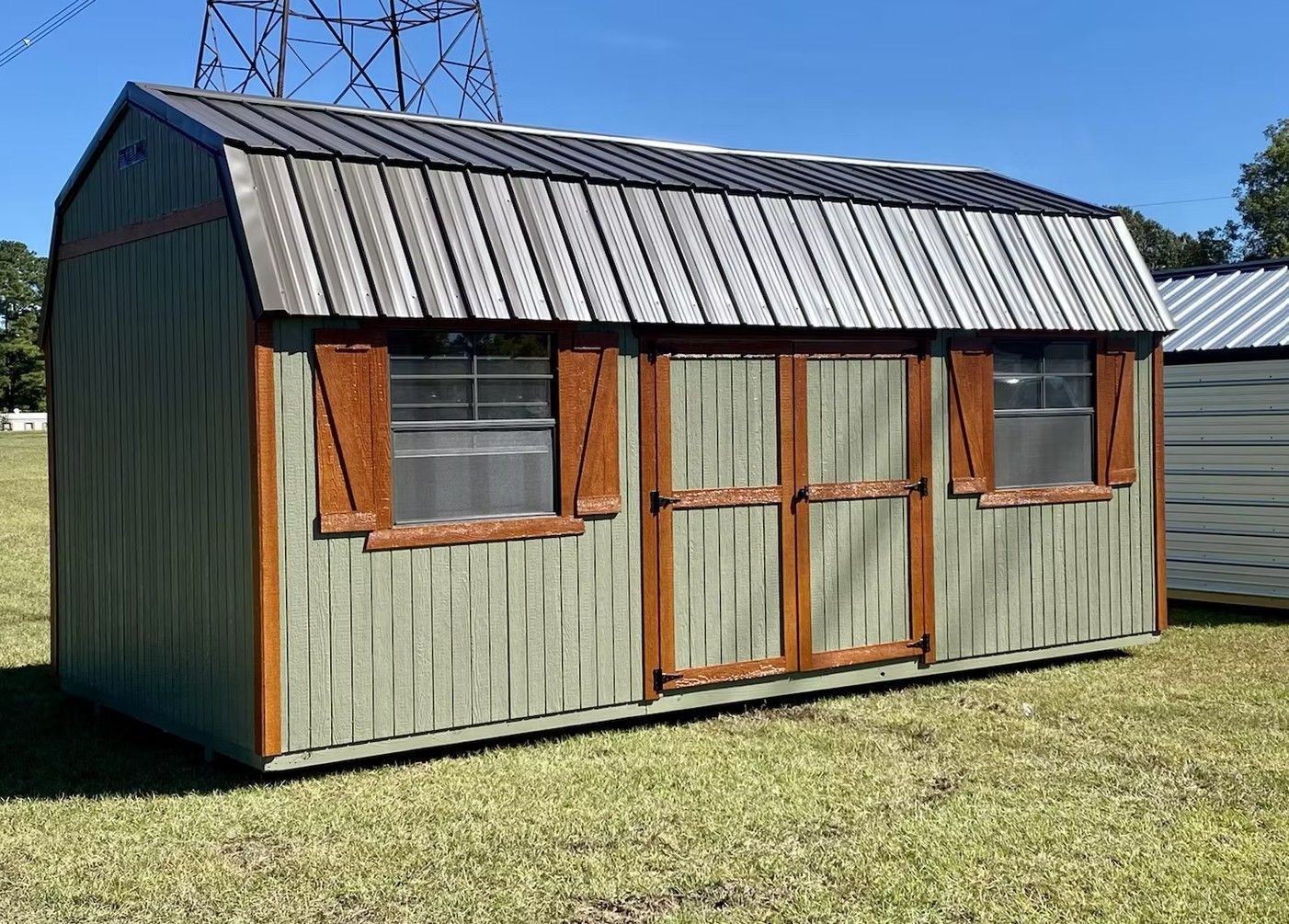 Green shed with brown trim, two windows, and metal roof, sitting on grass under a clear blue sky.