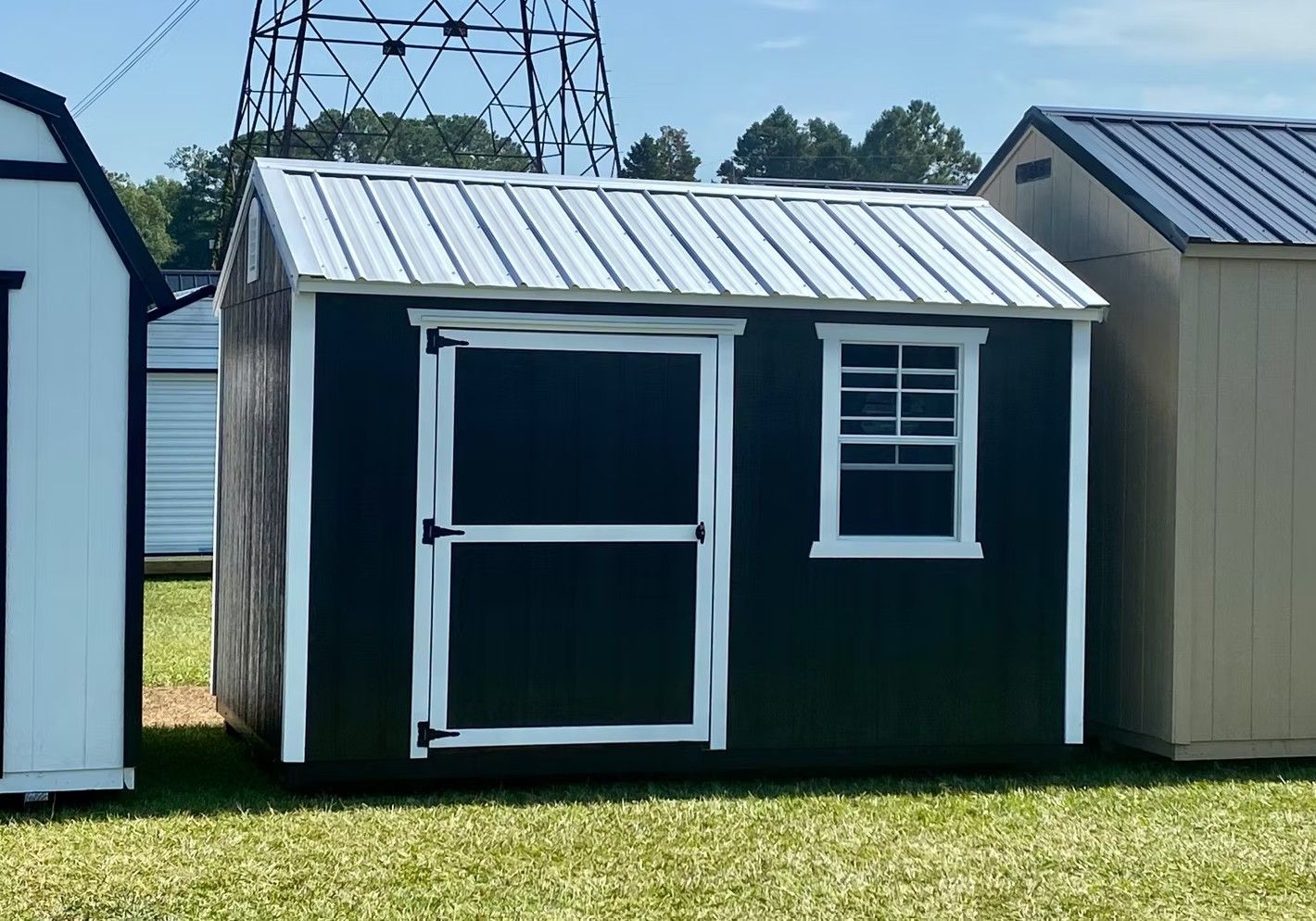 Dark green shed with white trim, door, and small window, on grass, other sheds in background.