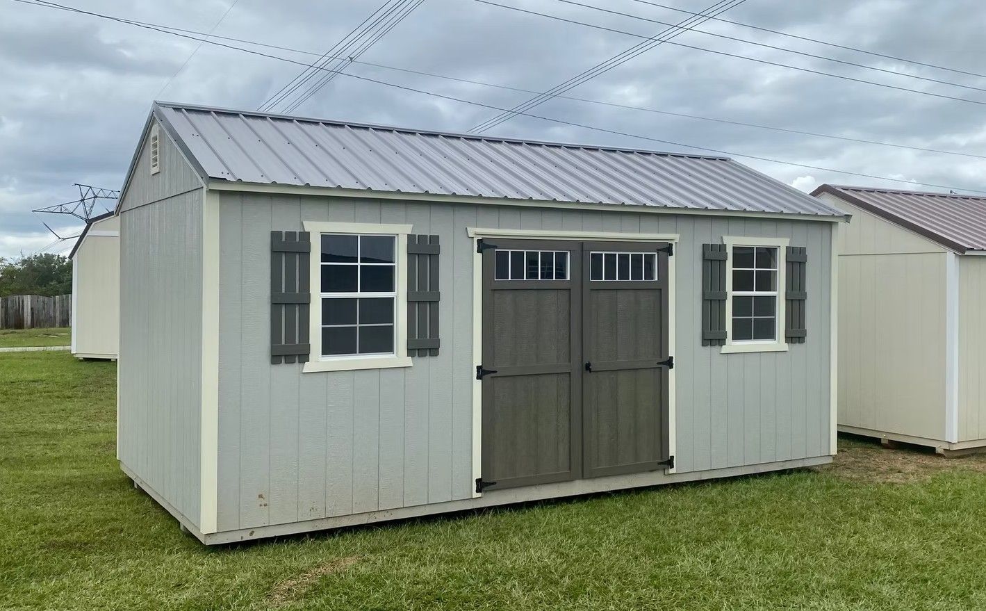Gray shed with metal roof, two windows with shutters, and double doors on a grassy field.