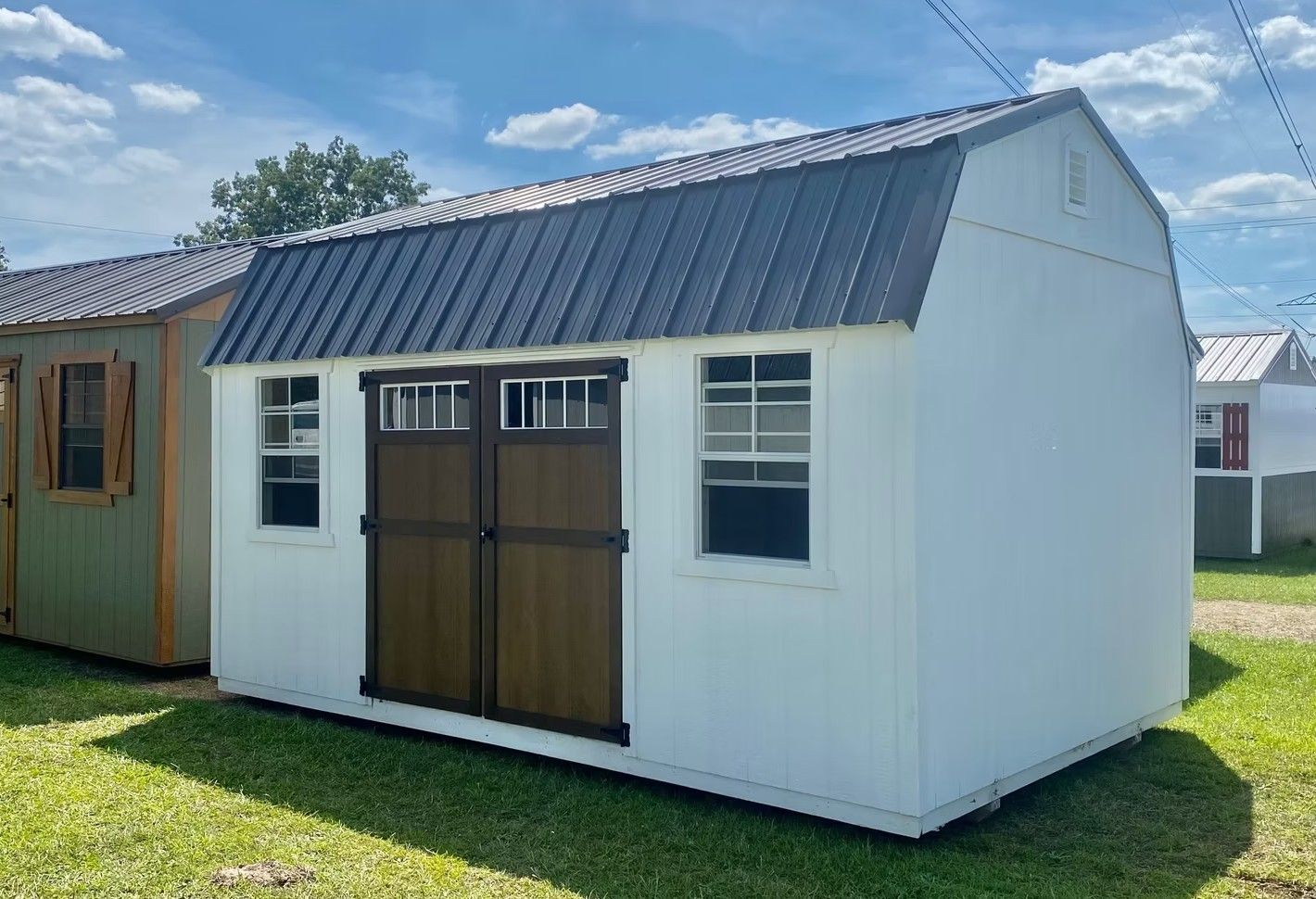 White shed with dark metal roof, brown doors, and two windows on a grassy lawn.