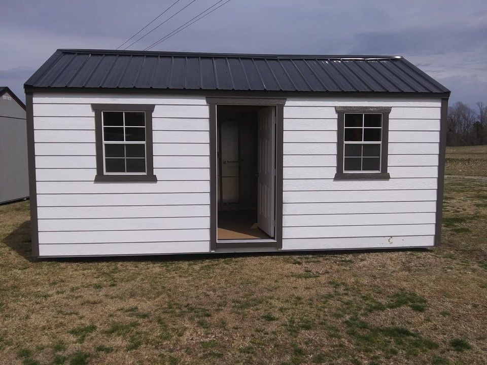 White shed with dark roof, open doorway, two windows. Brown trim. Located outdoors in grass.