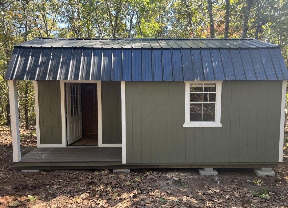Green shed with blue roof and white trim, porch with open door and window in a wooded area.