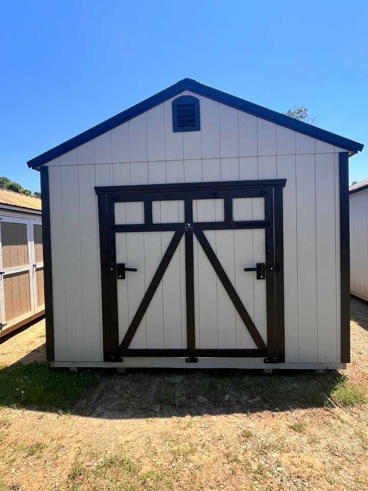 White shed with dark brown trim, double doors, and an air vent, set on grass under a blue sky.