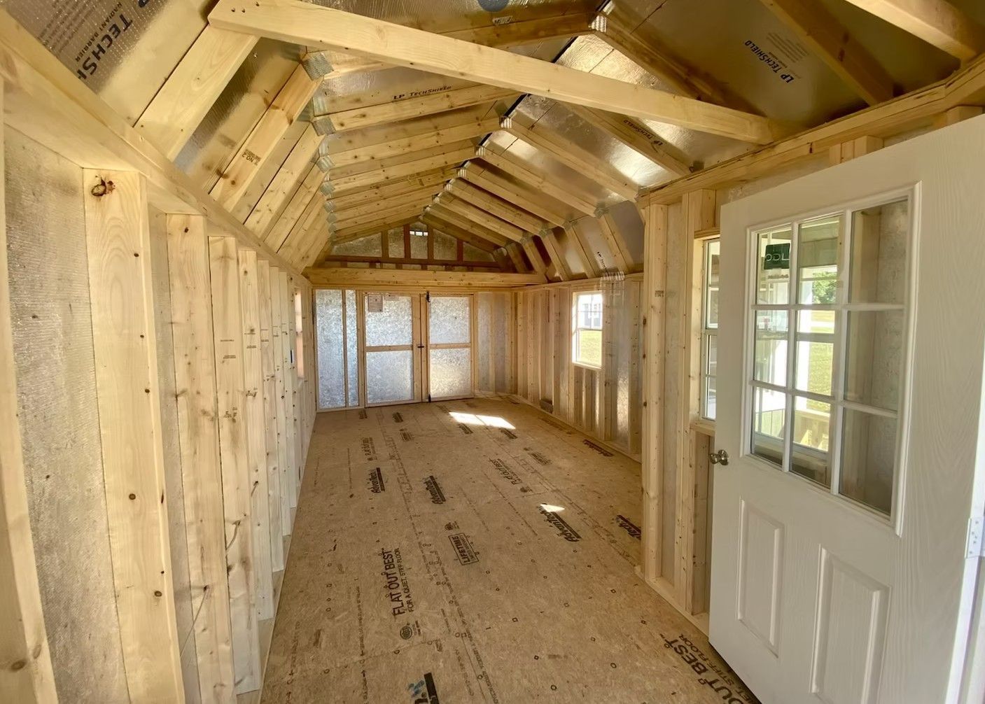 Interior of a wooden shed under construction, showing walls, floor, roof framing, and a door with windows.