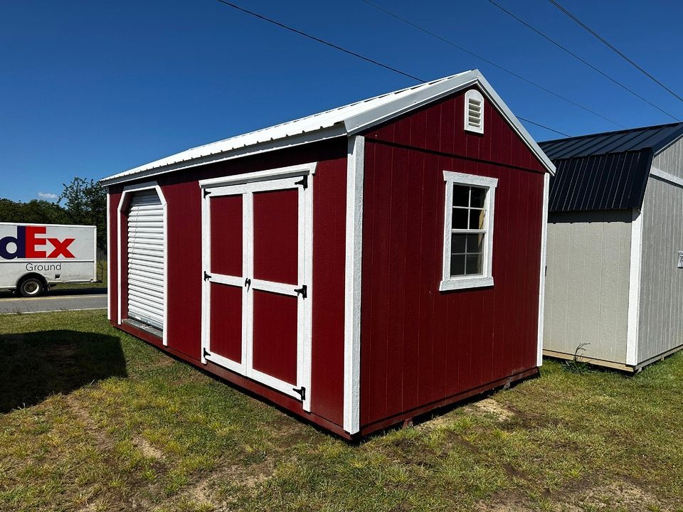 Red shed with white trim, a garage door, a window, and a white metal roof on display outdoors.