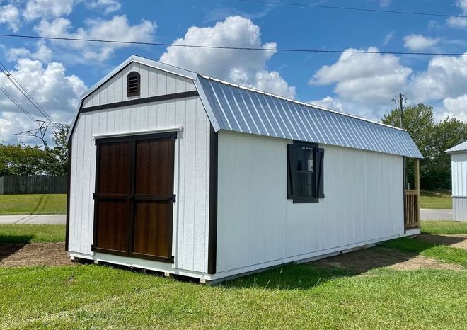 White barn-style shed with dark brown doors and trim, metal roof, and small window on grass under a cloudy sky.