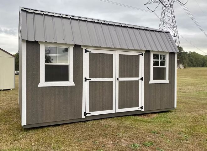 Gray shed with white trim, metal roof, double doors, and two windows in a grassy field.