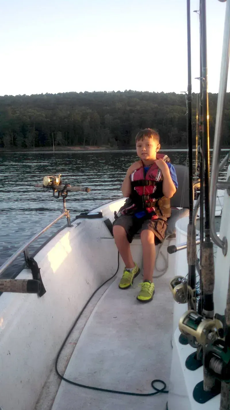 Person sitting on a sailboat deck wearing a pink life jacket, with calm water and forested shoreline behind.