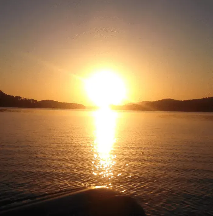 Sun setting over calm water, with golden reflections and silhouetted hills on the horizon