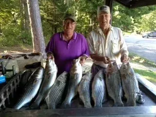 Two people holding large fish on a picnic table outdoors under a shaded shelter.