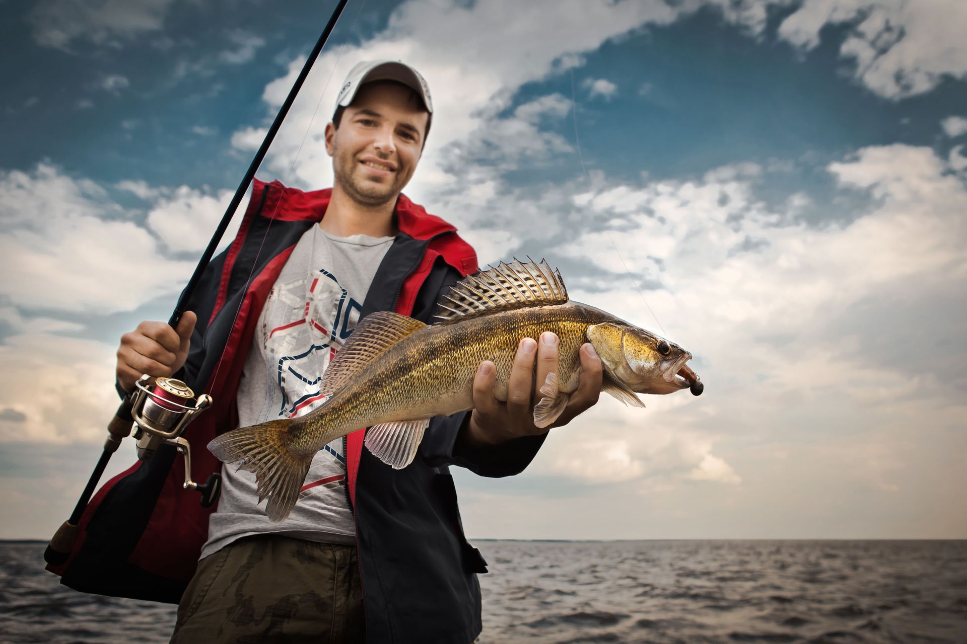 Angler holding a large fish on a boat, with a fishing rod and cloudy sky over the water.