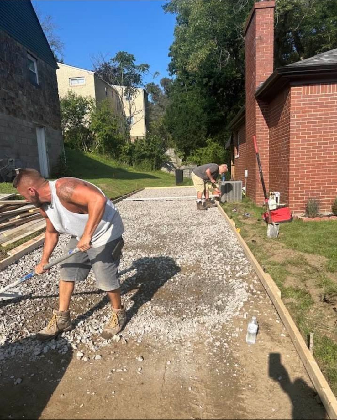 Two construction workers leveling gravel on a concrete patio next to a brick house. One is using a rake, the other a small wheelbarrow.