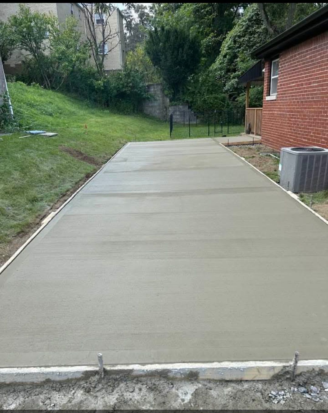 Newly poured concrete patio, level and gray, framed by forms. The background shows a grassy hill and a brick house.