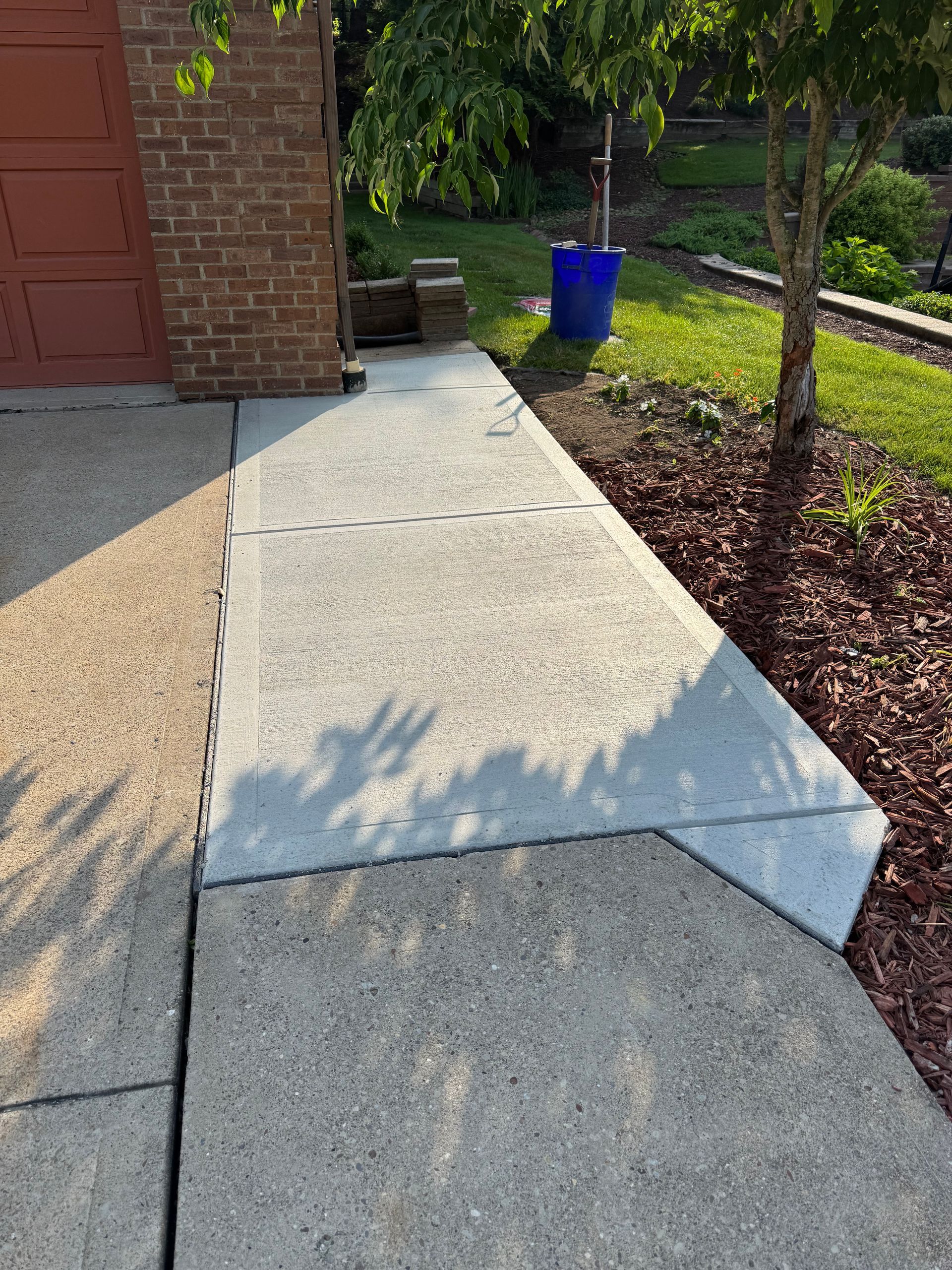 Concrete sidewalk with a sloped section leading to a garage, bordered by grass and a small tree.