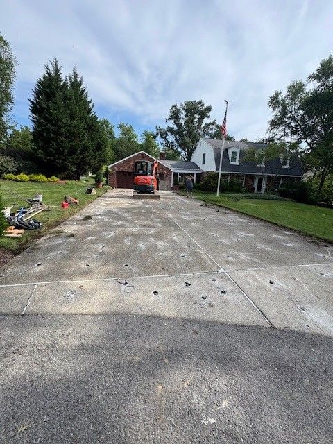 Driveway being demolished in front of a house with a red brick garage and a white house, with an excavator on the driveway.