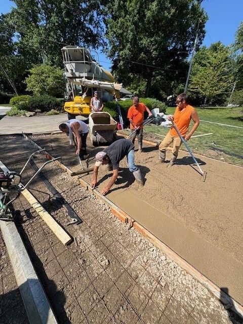 Construction workers pouring and leveling concrete for a driveway. Several men in orange shirts work near a concrete mixer, with tools and rebar visible.