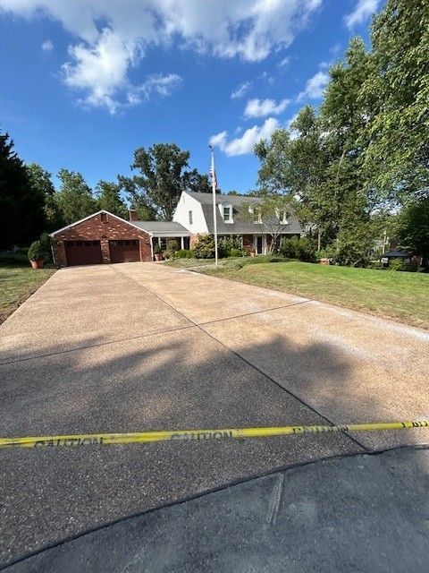 A house with a long concrete driveway under a blue sky, with caution tape in the foreground.  A brick garage sits to the left.