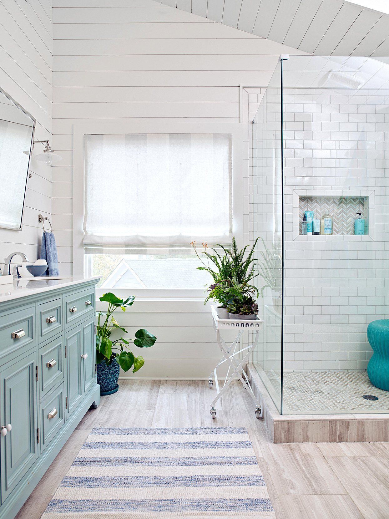 Coastal-style bathroom with light blue vanity, white walls, glass shower, and blue and white rug.