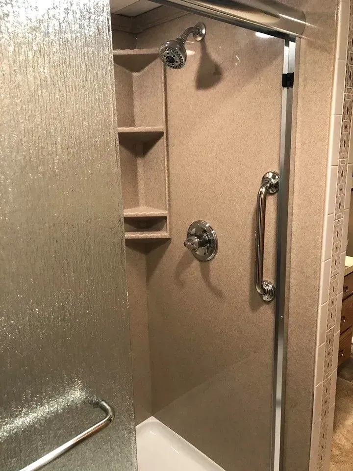 Shower stall with textured tan walls and glass door, featuring a showerhead, shelving, and a grab bar.