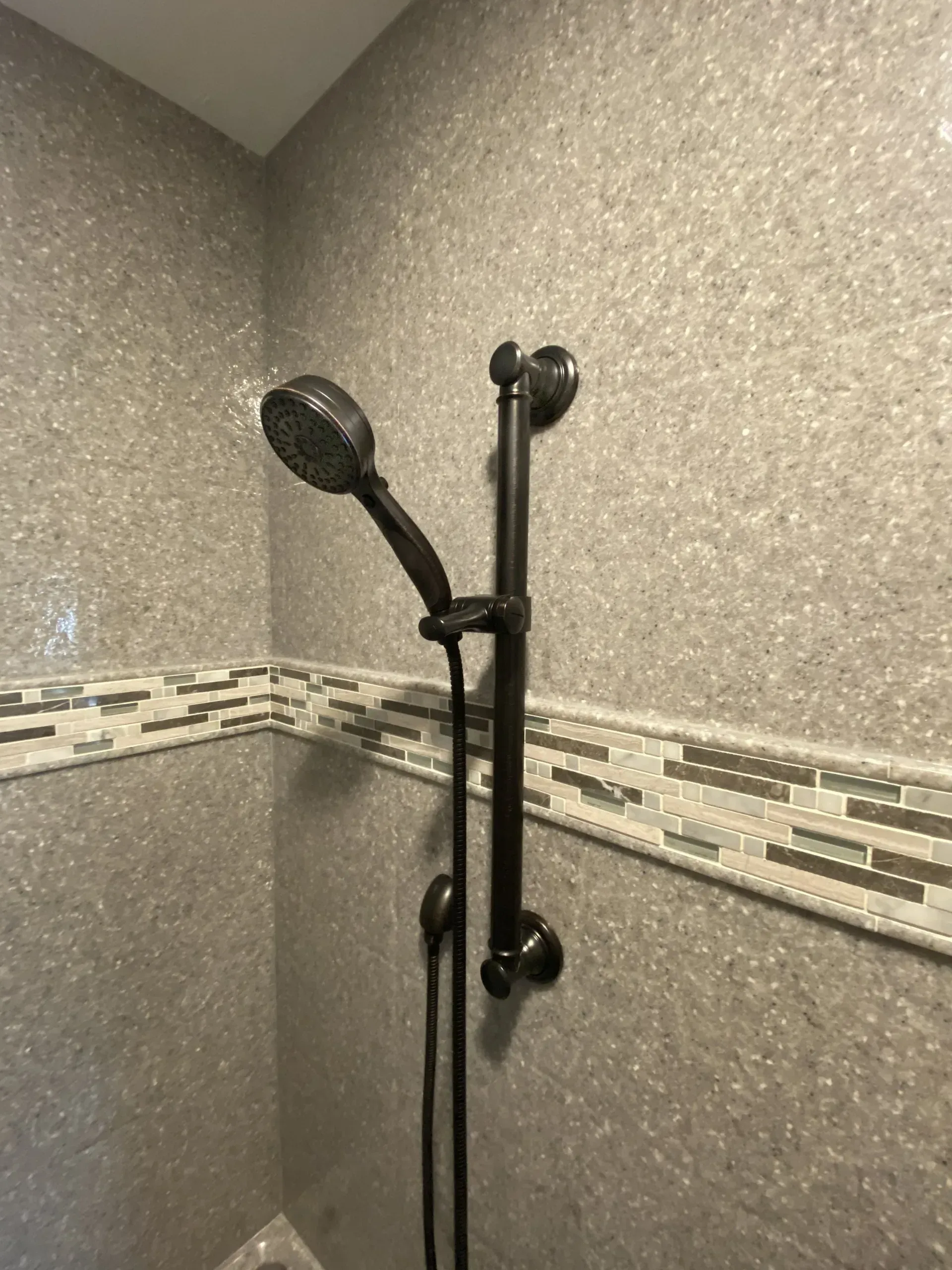 Black shower head on a dark vertical rail in a gray tiled shower with a decorative border.