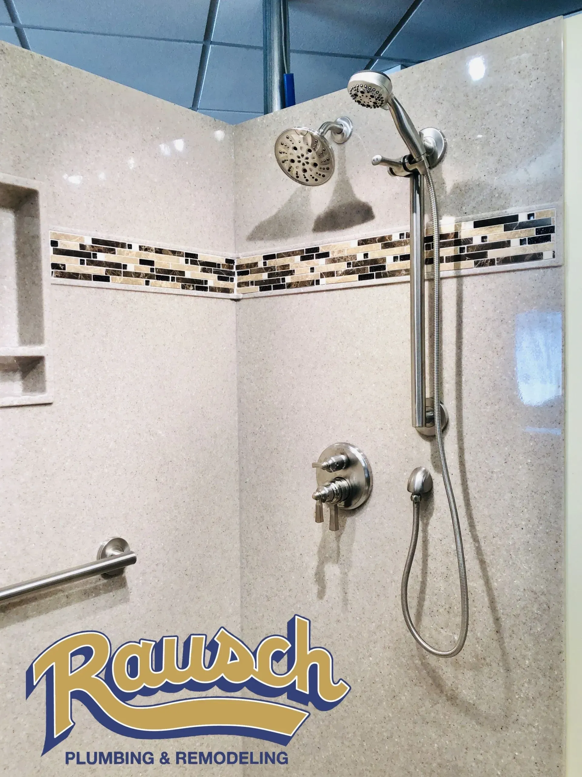 Corner shower with speckled gray walls, mosaic tile border, silver shower fixtures. Rausch Plumbing & Remodeling logo.