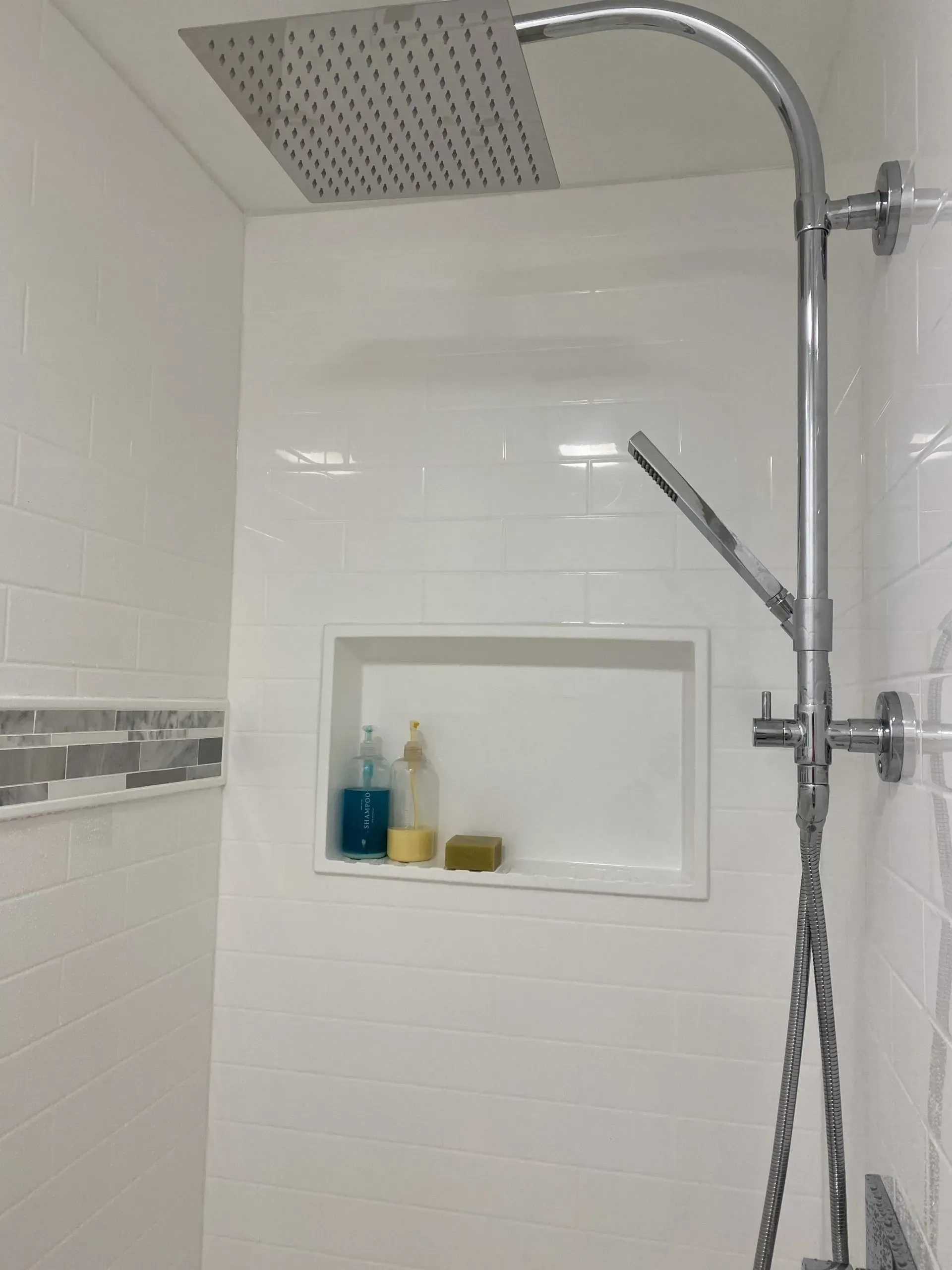 Modern white tiled shower with rain showerhead, handheld shower, and recessed shelf with soap and bottles.